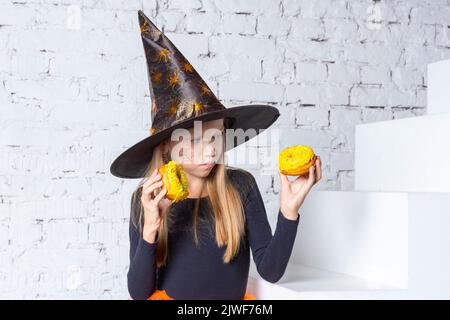 Halloween bambini. Una bambina bionda sorridente in costume da strega che tiene in mano le ciambelle gialle, seduto sui gradini della casa. Halloween felice Foto Stock