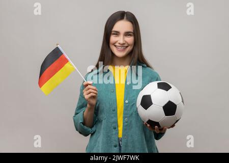 Donna sorridente soddisfatta che tiene in mano la bandiera della Germania e la sfera di calcio, che sostiene la squadra favorita sul campionato, che indossa la giacca casual di stile. Studio in interni isolato su sfondo grigio. Foto Stock