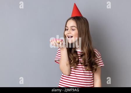 Ritratto di piccola ragazza gioiosa affamata che indossa T-shirt a righe e cono partito, tenendo e vuole mordere golosa ciambella dolce, guardando il dessert. Studio in interni isolato su sfondo grigio. Foto Stock