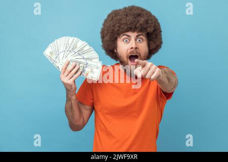 Ritratto di stupito scioccato uomo con acconciatura afro indossare T-shirt arancione in possesso di banconote in dollari, puntando alla fotocamera, si vince la lotteria. Studio in interni isolato su sfondo blu. Foto Stock