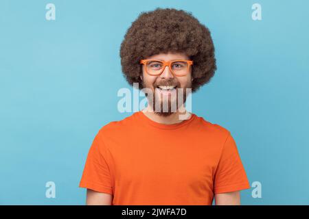 Ritratto di divertente uomo positivo con acconciatura afro con T-shirt arancione e occhiali, guardando la macchina fotografica con felice espressione e sorriso toothy. Studio in interni isolato su sfondo blu. Foto Stock