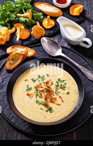 Kanttarellikeitto, zuppa finnica di Chanterelle in ciotola nera su tavolo di legno scuro con cucchiaio, funghi Chanterelle freschi sullo sfondo, vista verticale fr Foto Stock