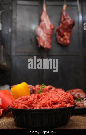 Vista laterale di manzo fresco sminuzzato e carni assortite sul tavolo rustico con tagli appesi sullo sfondo Foto Stock
