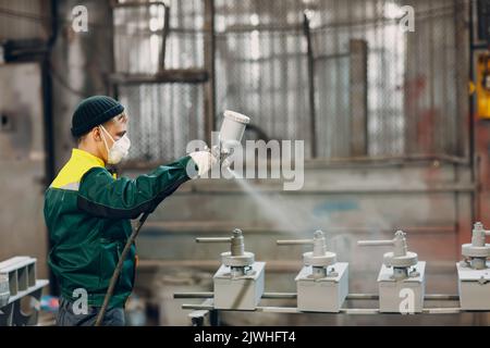 Rivestimento con primer per polveri di parti metalliche. L'uomo di lavoro in un vestito protettivo spruzza la vernice in polvere dalla pistola sulla costruzione del prodotto metallico nello stabilimento di fabbrica. Foto Stock