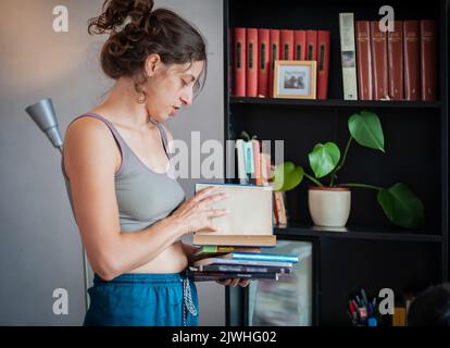 Una giovane donna rilassata mette diversi tipi di libri su uno scaffale nella stanza del suo appartamento Foto Stock
