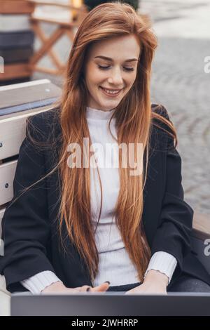 Giovane donna rossa sorridente seduta sulla panchina del parco navigando sul web con il suo laptop. Rete Internet. Stile di vita cittadino. Cura dei capelli. Foto Stock
