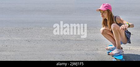 Ritratto di una ragazza caucasica sorridente con capelli lunghi in un cappellino rosa da baseball, seduta su uno skateboard su una strada asfaltata in condizioni di sole. Un bambino cavalca Foto Stock