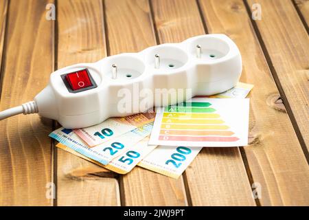 Presa elettrica, etichetta energetica e moneta in euro sul tavolo di legno. Concetto di aumento dei prezzi elettrici. Foto Stock