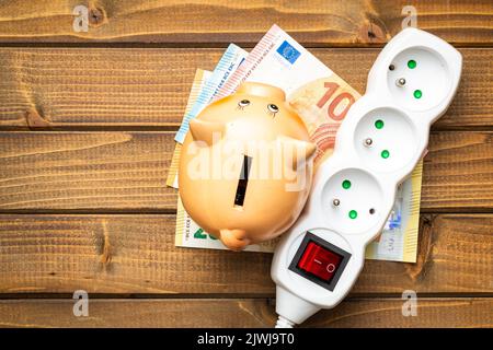 Presa elettrica, banco di porcellini e denaro in euro sul tavolo di legno. Concetto di aumento dei prezzi elettrici. Vista dall'alto. Foto Stock