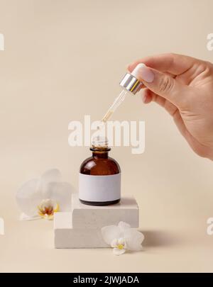 Tenere la pipetta sopra il flacone contagocce cosmetico aperto vicino a fiori di orchidea bianca e pietre su beige chiaro, primo piano, mockup. Produ naturale per la cura della pelle Foto Stock