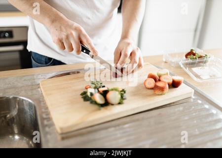 Un uomo che indossa una t-shirt bianca che taglia le fragole in una cucina su un asse di legno con un coltello Foto Stock