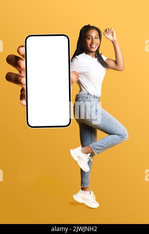 Donna allegra afroamericana che salta e mostra enorme smartphone con schermo vuoto, felice ragazza nera millenaria pubblicità qualcosa, consiglia nuova app mobile Foto Stock