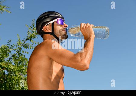 Il triatleta beve acqua dalla sua bottiglia con gli occhiali da sole e senza camicia. Foto Stock