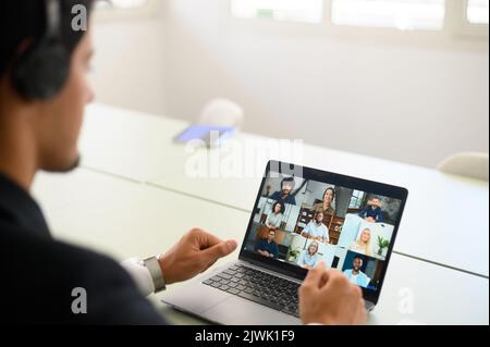 Uomo d'affari latino che utilizza il laptop per la comunicazione video con diversi team di lavoro internazionali, parlando a distanza con un gruppo di colleghi multirazziali, vista posteriore sullo schermo del computer. Videochiamata Foto Stock