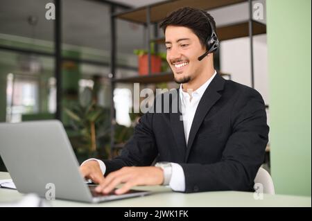 Allegro dipendente di un ufficio con cuffie che utilizza un notebook seduto sul posto di lavoro, videochattare con il datore di lavoro o i clienti, uomo d'affari che utilizza cuffie con microfono per le comunicazioni online con i clienti Foto Stock