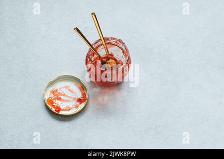 Marmellata di fragole dolci mangiata per due. Vasetto in vetro con avanzi confiture, coperchio e due cucchiai su fondo grigio. Foto Stock