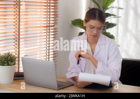 Giovane donna dottore che usa un computer portatile e che guarda un documento sul paziente per la diagnosi in clinica, medico femminile di lavoro e di lettura docum Foto Stock