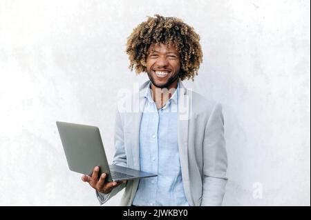 Ritratto di bel positivo successo ricci capelli misti razza uomo d'affari, elegantemente vestito, tenendo in mano il portatile aperto, si alza su sfondo isolato, guarda la fotocamera, sorridendo amichevole Foto Stock