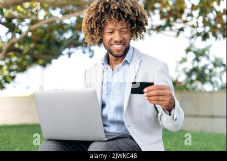 Positivo bel latino o brasiliano uomo, sedersi all'aperto, utilizzando una carta di credito e un computer portatile, fare acquisti online, effettuare pagamenti, ordinare la consegna di cibo, inserire i dati, sorrisi. Acquisti online, pagamento Foto Stock