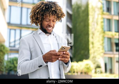 Scrivere un messaggio, navigare nei social media. Positivo bello fiducioso riccio capelli misti uomo di razza, imprenditore di successo, in piedi all'aperto, utilizzando smartphone, messaggi nei social network, sorridente Foto Stock