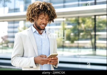 Messaggistica online, navigazione in Internet. Positivo misto razza uomo con capelli ricci, indossando abiti eleganti formale, si trova all'aperto vicino al business center, utilizza il suo cellulare, guarda lo schermo del dispositivo, sorride Foto Stock