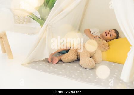 Concetto di infanzia moderna. Adorabile ragazzo europeo dai capelli castani vestito casualmente pronto a fare un pisolino con il suo orsacchiotto beige in una comoda tenda tipi di colore chiaro posta nel soggiorno. Foto di alta qualità Foto Stock