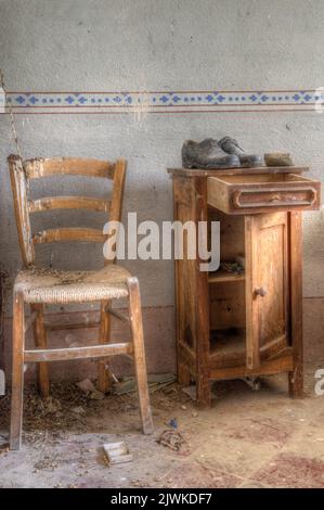 Italia, 09-10-2023. Vecchia sedia e comodino con un paio di scarpe decadenti. In una vecchia casa di campagna abbandonata nel nord Italia Foto Stock