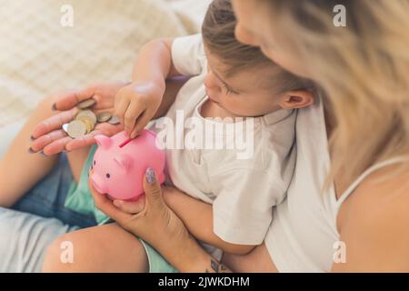 Madre che si lega al figlio. Giovane donna incinta caucasica bionda insegnando al figlio di quattro anni come risparmiare denaro utilizzando piggybank rosa. Ragazzo mettere monete. Foto di alta qualità Foto Stock