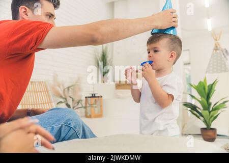 Quattro feste di compleanno. Orgoglioso commesso papà caucasico mettere cappello partito sulla testa di suo figlio. Il ragazzino che cerca di far esplodere il corno. Foto di alta qualità Foto Stock