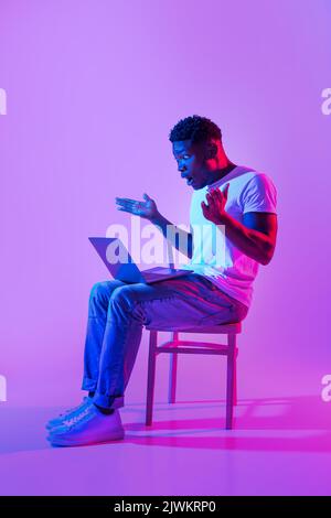 Emotivo giovane uomo nero seduto sulla sedia con notebook, sensazione di scioccata, facendo errore nel progetto di business in luce al neon Foto Stock