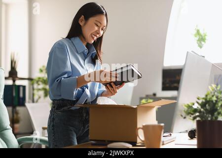 Allegro Asian Businesswoman Unpacking scatola Holding Book in piedi in ufficio Foto Stock