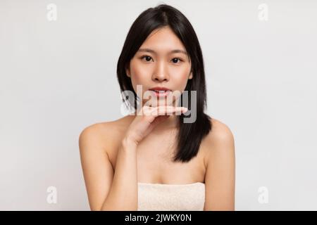 Bellezza naturale. Ritratto di giovane donna asiatica avvolto in un asciugamano che tocca il mento e guarda la macchina fotografica, sfondo bianco Foto Stock