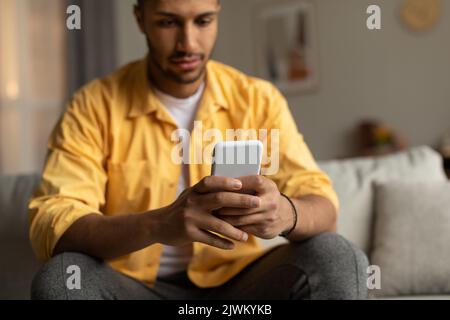 Giovane ragazzo afro-americano seduto sul divano con lo smartphone, chattare online, navigare sul social network a casa Foto Stock