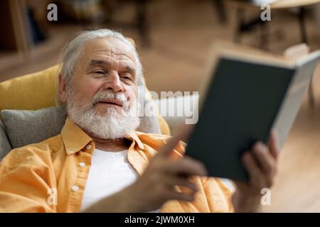 Uomo anziano positivo che si posa sul divano, leggendo il libro Foto Stock