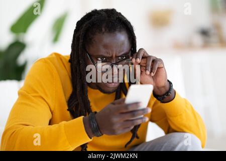 Concetto di problemi di vista. Giovane uomo nero che indossa occhiali lettura messaggio sullo smartphone Foto Stock