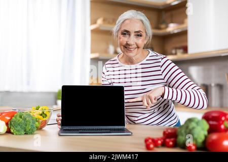 Donna anziano felice che indica lo schermo vuoto del laptop in cucina Foto Stock