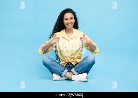 Happy Arabic Female gesturing Thumbs Up seduto su sfondo blu Foto Stock