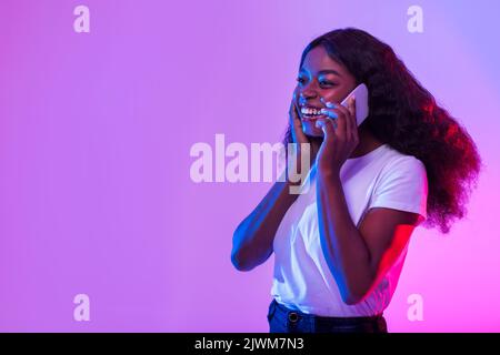 Affascinante giovane donna nera che parla sul cellulare, avendo conversazione telefonica in luce al neon, spazio vuoto Foto Stock