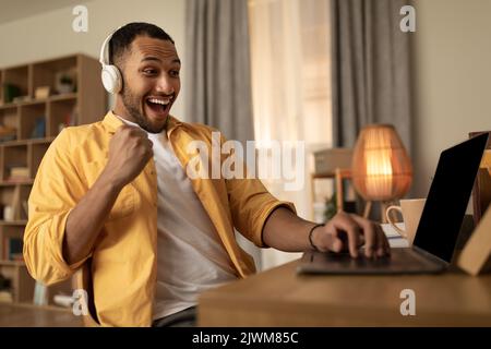 Giovane uomo nero eccitato in cuffie gesturing sì, festeggiando il successo o grande vittoria, seduto vicino al PC portatile a casa Foto Stock