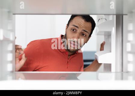 Disperato triste scioccato giovane afroamericano ragazzo apre porta sguardi di frigorifero, controlla cibo o ghiaccio in caldo giorno Foto Stock
