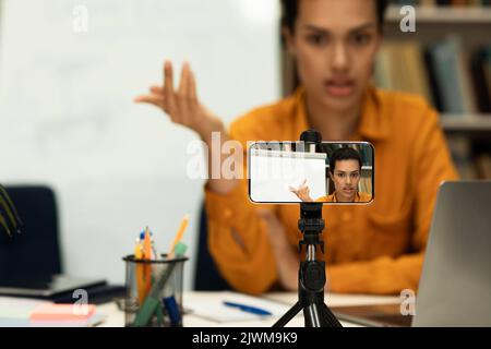 Giovane insegnante di sesso femminile che fornisce lezioni online guardando la fotocamera del cellulare o la registrazione di video su smartphone su treppiede Foto Stock