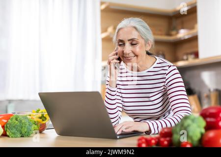 Bella donna anziana che parla sul cellulare e utilizzando il computer portatile in cucina Foto Stock