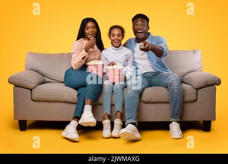 La famiglia afro-americana era molto felice di guardare film e mangiare popcorn dai secchi, i genitori seduti con la figlia sul divano Foto Stock