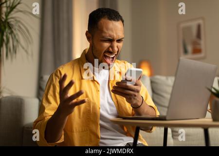 Concetto di stress lavorativo. Un giovane nero arrabbiato che urla nello smartphone, lavorando online sul PC portatile in ufficio Foto Stock