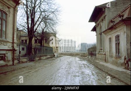 Bucarest, Romania, gennaio 1990, poche settimane dopo la caduta del comunismo. Mentre Ceausescu si concentrava sulla costruzione dei suoi progetti megalomanici, come il Centro Civico, i quartieri storici della capitale si deterioravano lentamente. Alcune case storiche nelle vicinanze del Blvd. Della Vittoria del socialismo sono state evacuate per demolizione, e a malapena sfuggite grazie alla Rivoluzione anticomunista. In questa foto, una strada fangosa nel vecchio quartiere '11 Iunie', con una vista dei nuovi imponenti edifici lungo il Boulevard della Vittoria del socialismo. Foto Stock
