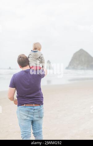 Il bambino biondo, caucasico, siede sulle spalle del padre sulla spiaggia di Cannon. Foto Stock