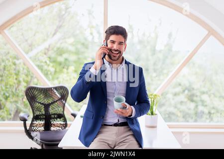 Buon uomo d'affari che effettua una telefonata e ride mentre si trova in ufficio. Indossa una camicia e un blazer. Foto Stock