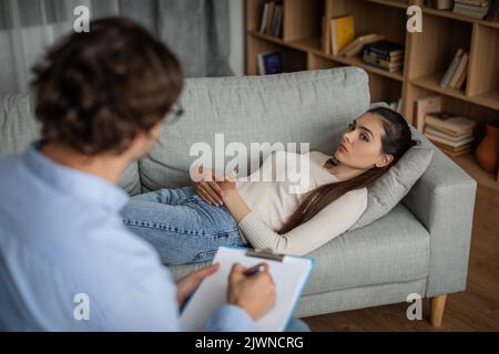 Triste donna europea millenaria che soffre di problemi mentali si trova sul divano, parlare con l'uomo medico in clinica Foto Stock