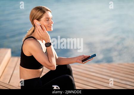 Sorridente Donna di mezza età negli auricolari che riposano con lo smartphone dopo l'allenamento all'aperto Foto Stock