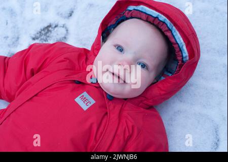 Ritratto di un bambino di nove mesi in un manto di neve rosso sdraiato sulla neve in un giorno d’inverno. Foto Stock